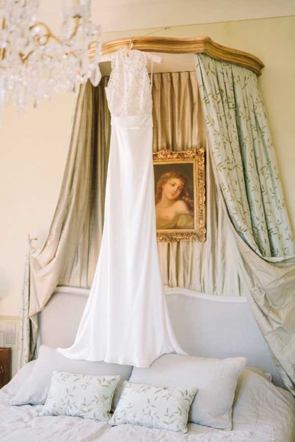A getting-ready detail shot of a white wedding gown hanging from the gilded canopy frame of a four-poster bed. The dress features a lace bodice with a sleeveless, high-neck design and a simple, flowing crepe skirt. The bed has sage green silk canopy drapes with an embroidered floral motif and pale blue embroidered cushions on a white bedspread. A gold-framed oil portrait painting hangs on the wall behind the headboard, and a crystal chandelier is partially visible in the upper left corner. The overall room styling is classic French château with an ornate, period-appropriate interior.
