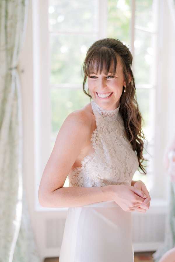 A getting-ready portrait of a bride standing in front of large French windows with sheer white curtains, allowing bright natural light to fill the indoor space. The bride is wearing an ivory lace halter-neck wedding dress and has her dark brown hair styled in a half-up, half-down look with soft waves and bangs. She is smiling broadly while someone just out of frame appears to be fastening or adjusting the back of her dress. The shot is a close-up portrait with a softly blurred background.