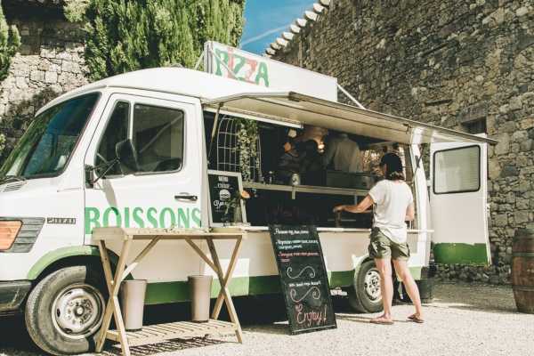 A white and green food truck labeled 'PIZZA' and 'BOISSONS' is set up outdoors against a stone wall, serving guests during what appears to be a wedding cocktail hour or reception. A wooden folding table is positioned in front of the truck, and a chalkboard A-frame sign reading 'Enjoy!' is placed on the gravel ground nearby. One person in a white t-shirt and shorts is standing at the service window, and several people are visible inside the truck. The setting is a rustic outdoor venue with stone architecture, and the image is a medium wide shot taken in bright natural daylight.