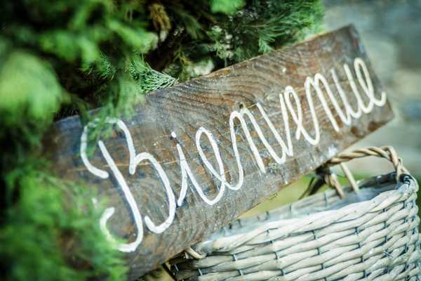 Close-up detail shot of a rustic wooden sign with 'Bienvenue' written in white hand-lettered script, propped inside a woven wicker basket with greenery visible in the background. The sign is made from a raw, dark-stained wood plank, consistent with a rustic or bohemian wedding styling theme. The shallow depth of field keeps the sign sharp while the background greenery is softly blurred.