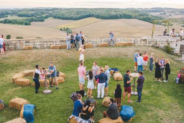 An outdoor cocktail hour taking place on a hilltop terrace or lawn with panoramic views of rolling agricultural fields in the background. Approximately 30-40 guests are mingling in small groups across the space. The seating and lounge areas are styled with hay bales arranged in clusters, some topped with blue fabric cushions, alongside tall cocktail tables with metal stands — a relaxed rustic theme. Guests are dressed in casual summer attire including bright colors and shorts, suggesting an informal dress code. The shot is taken from an elevated angle, likely from a balcony or upper floor, giving a wide overhead perspective of the entire cocktail area.