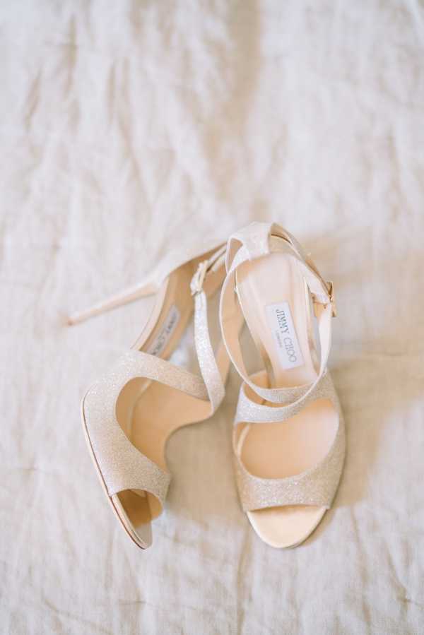 A detail close-up flat lay of a pair of Jimmy Choo bridal heels placed on a white linen fabric surface. The sandals feature a champagne gold glitter finish, open toe, and ankle strap with a gold buckle closure. The stiletto heel is slender and the insole lining is nude/blush tone. The Jimmy Choo brand label is visible on the inner lining of the right shoe.