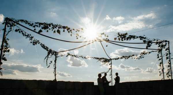 A wide-shot silhouette of two people — appearing to be a couple — standing beneath an outdoor ceremony arch or chuppah structure draped with trailing green vine foliage and rope or fabric swags, with a third person (likely an officiant) facing them. The image is strongly backlit by a bright sunburst low in the sky, rendering all figures and the arch as dark silhouettes against a blue sky with scattered clouds. The ceremony is set outdoors, positioned against what appears to be a low stone wall. The overall styling is natural and boho, with the greenery-draped structure as the primary decor element.