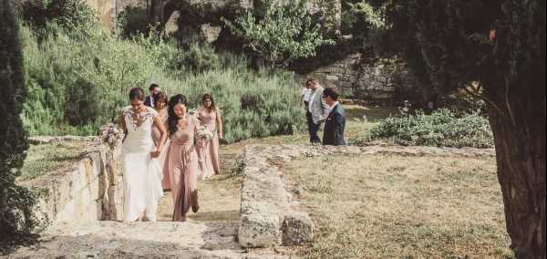 The bridal party is walking up a set of rustic stone steps in an outdoor garden setting, likely at a historic chateau or domaine. The bride wears a fitted white gown with lace detailing on the bodice, accompanied by three bridesmaids in floor-length blush pink dresses carrying small bouquets. Two male guests or groomsmen in suits are visible in the background to the right. The wide shot captures the full bridal party in motion, framed by mature trees and old stone walls, with a natural, relaxed feel consistent with a rustic or classic French countryside aesthetic.