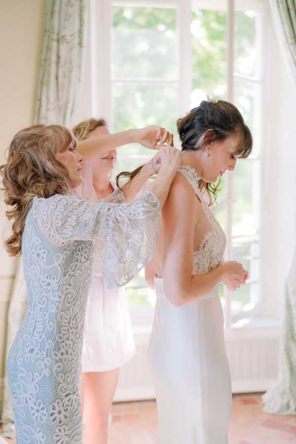 A getting-ready scene photographed indoors, with two women helping a bride into her wedding dress in front of large French windows with sheer white and sage green curtains. The bride has dark hair styled in an updo with a small crystal hairpiece and is wearing a fitted ivory gown with a lace bodice and low open back. One helper wears a sage blue lace dress with wide flutter sleeves, and the other wears a short white dress; both appear to be fastening the back of the bride's gown. The shot is a medium portrait taken from a slight side angle, with soft natural backlight streaming through the windows.