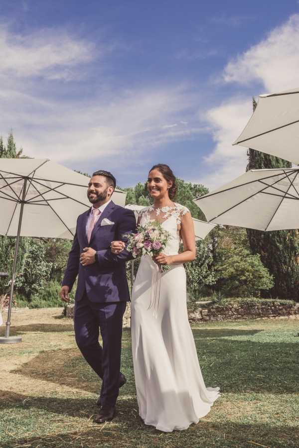 A bride and groom walk arm-in-arm through an outdoor garden setting, smiling broadly in what appears to be a post-ceremony recessional or garden portrait moment. The groom wears a navy blue suit with a pink tie and white pocket square, while the bride wears a fitted ivory gown with a lace illusion cap-sleeve bodice and a flowing skirt with a short train. The bride carries a bouquet of mauve and lilac roses with soft pink blooms and greenery. Large white market umbrellas flank the couple on both sides, and the setting features a landscaped garden with stone boundary walls in the background. The composition is a full-length portrait shot taken at ground level with natural daylight.