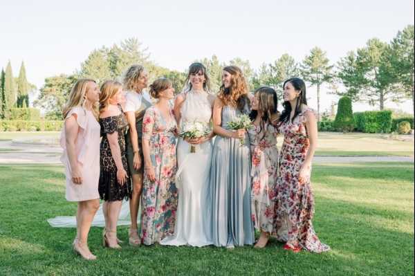 A bridal party portrait taken outdoors on a manicured lawn, likely at a garden or chateau venue, in warm evening light. The bride stands at the center wearing a fitted ivory halter-neck gown and holding a small bouquet of white and cream flowers with greenery, surrounded by seven bridesmaids or female guests. The group wears a mix of mismatched dresses including floral print maxi dresses in red, pink, and green tones, a pale blush mini dress, a black lace dress, and a floor-length powder blue wrap-style dress, reflecting a relaxed, bohemian styling approach. The women are gathered closely together, laughing and looking toward the bride in a candid, conversational pose. The shot is a medium-wide portrait at eye level.