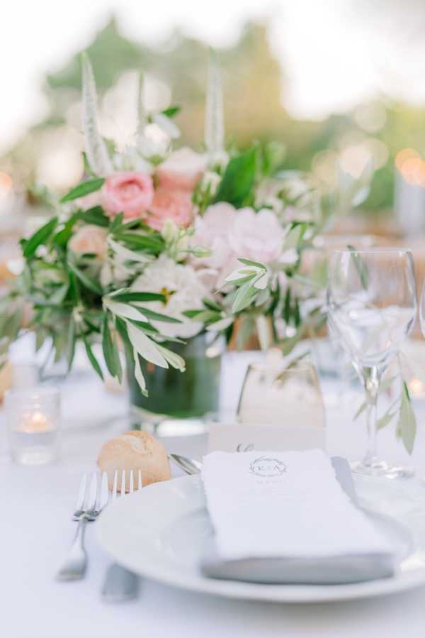 A close-up detail shot of an outdoor wedding reception table setting with a white linen tablecloth. The centerpiece features a low arrangement of blush pink roses, white flowers, and olive branch foliage in a dark green glass vessel, flanked by small votive candles and a bud vase with a single olive sprig. The place setting includes a white plate with a white folded napkin bearing a monogrammed crest logo, silver fork and knife, multiple wine glasses, and a small bread roll. The overall decor palette is white, blush pink, and soft green, consistent with a classic, garden-inspired style.