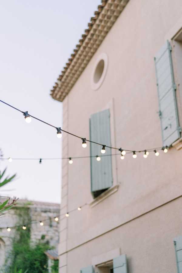 A detail shot taken at dusk showing strands of globe string lights suspended outdoors in front of a classic Provençal stone building with pale terracotta-toned render, sage green shutters, a circular oculus window, and terracotta roof tiles. No people are visible; the image focuses on the warm-glowing bulbs strung diagonally across the frame as an outdoor lighting decor element. The composition is a low-angle, close-up shot with the building softly out of focus in the background. Potential venue feature image.