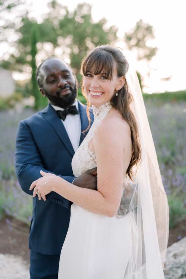A couple portrait shot outdoors, likely taken during golden hour based on the warm light. The bride wears a white halter-neck gown with a lace bodice and a long flowing veil, with her dark hair worn half-up with bangs. The groom stands behind her with his arms around her waist, wearing a navy blue suit with a black bow tie. The background is softly blurred but shows rows of purple lavender in bloom, suggesting a Provençal setting. The composition is a medium close-up portrait with the bride turned slightly toward the camera and smiling directly at it.