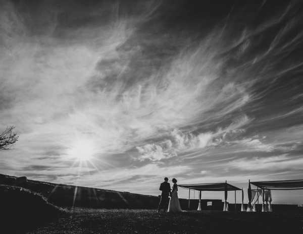 A black-and-white outdoor couple portrait shot at sunset, with the couple rendered as dark silhouettes against a bright sun and dramatic wispy cloud formations filling most of the frame. The couple, identifiable as a bride in a floor-length gown and a groom in a suit, stand close together facing each other in the lower left portion of the image. To the right, an open-sided tent or pergola structure with draped white fabric panels is also visible as a silhouette. The wide-angle composition emphasizes the expansive sky over the landscape, with high contrast between the dark foreground elements and the luminous backlit sky.