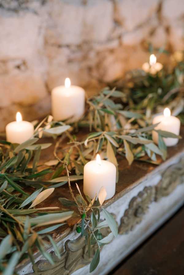 Close-up detail shot of a wedding decor arrangement featuring five lit white pillar candles interspersed with trailing olive branch garlands, including small green olives still on the stems. The arrangement is displayed on an ornate carved stone or plaster trough with decorative relief molding, positioned against a rough stone wall backdrop. The styling is rustic and Mediterranean in feel, with a warm candlelit glow and a natural green and white palette. No people are present in the frame.