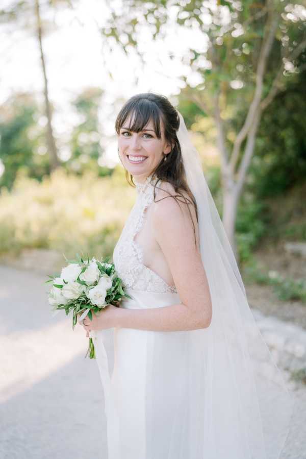 A bridal portrait taken outdoors on a gravel path with softly blurred greenery in the background. The bride, a dark-haired woman with bangs and a half-up hairstyle, faces slightly away from the camera and turns her head back toward the lens with a smile. She wears a white halter-neck gown with a lace bodice featuring a V-neckline and a flowing skirt, paired with a long cathedral-length veil draped over one shoulder. She holds a compact round bouquet of white roses and ranunculus with green foliage. The composition is a three-quarter portrait shot with soft natural light and a shallow depth of field.