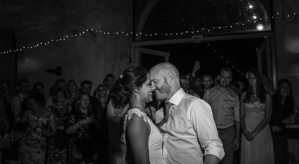 A black-and-white image of a couple sharing their first dance in what appears to be a rustic indoor venue, likely a barn or stone building. The bride wears a lace-detailed dress and has her hair up, while the groom is in a dress shirt with the collar open. The two are close together, smiling at each other, with a large crowd of approximately 20–30 guests forming a circle around them in the background. Fairy lights are strung along the ceiling, adding to the festive atmosphere of the reception. The image is a mid-range portrait shot with soft contrast typical of nighttime indoor photography.