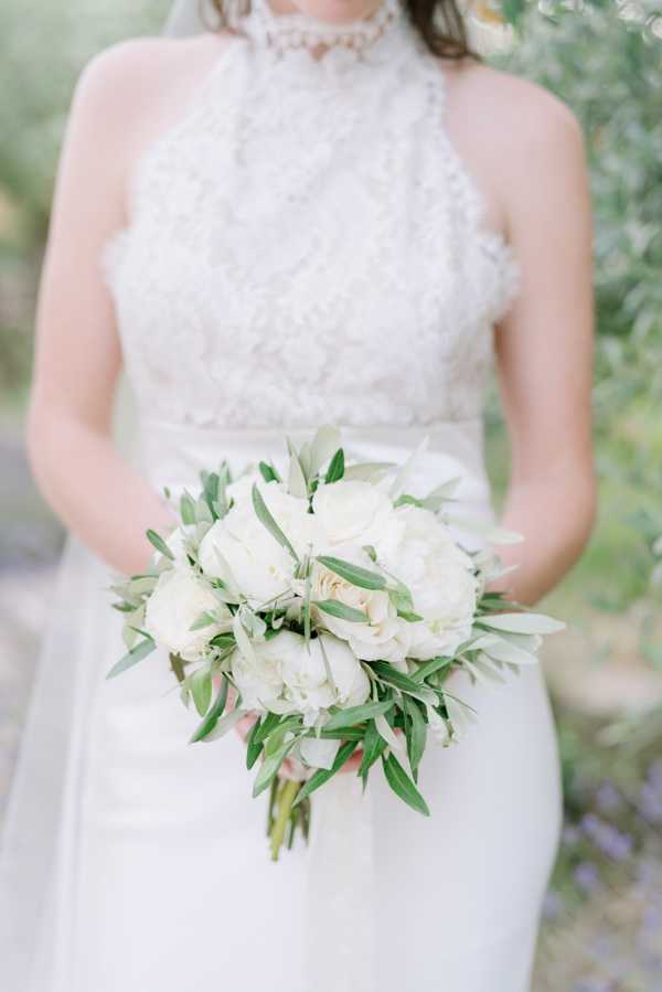 A close-up portrait of a bride holding her bouquet outdoors, with the background softly blurred. The bride wears a white halter-neck gown featuring intricate lace detailing on the bodice and a pearl or beaded choker necklace. The bouquet is a rounded arrangement of ivory peonies, white roses, and olive branch foliage, giving it a Mediterranean or Provençal feel. The composition is a mid-shot focused on the torso and bouquet, with a clean, light and airy photographic style.