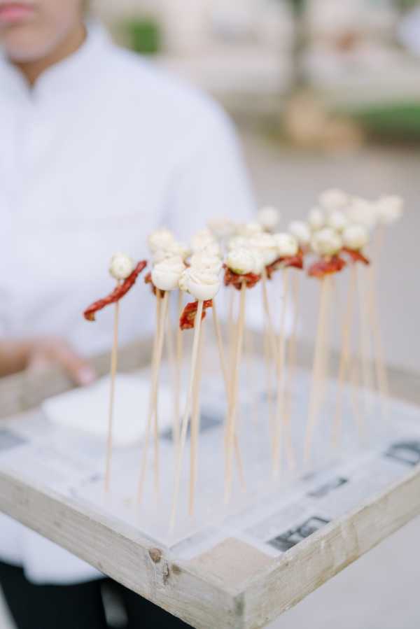 Close-up detail shot of cocktail hour canapés being passed by a server dressed in white. The appetizers consist of small cream cheese or whipped cheese rosettes paired with dried or semi-dried tomatoes, each presented on a tall wooden skewer. The skewers are displayed upright in a rustic whitewashed wooden tray lined with newspaper, creating a casual yet intentional presentation style. The server holding the tray is softly blurred in the background, keeping full focus on the food display.