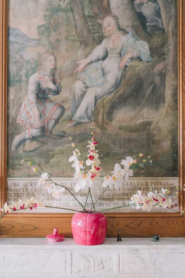 A close-up detail shot of a floral arrangement placed on a marble mantelpiece in front of a large antique painted panel or tapestry with French text inscription along the lower border. The arrangement features white and pink-centered phalaenopsis orchids with delicate trailing stems, displayed in a rounded hot-pink glazed ceramic vase. A small matching pink lidded ceramic dish sits beside it, along with two small dark cone-shaped objects and a tiny decorative piece on the marble surface. The painted panel behind depicts classical figurative scenes with soft muted tones in a French château interior style. The overall decor palette combines hot pink, white, and antique warm wood tones. Potential venue feature image.