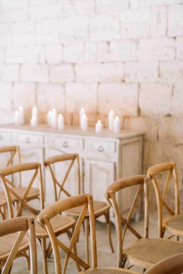 Detail shot of an indoor ceremony setup inside a whitewashed stone-walled venue, likely a French chateau or barn. Rows of natural wood cross-back chairs with woven rattan seats are arranged facing a distressed, pale grey painted sideboard or console table topped with multiple white pillar candles of varying heights. The decor palette is entirely neutral — warm honey-toned wood, off-white candles, and aged white paintwork — creating a minimal, rustic-classic aesthetic. The composition is a shallow depth-of-field portrait-orientation detail shot with the foreground chairs in soft focus and the candlelit sideboard as the focal point.