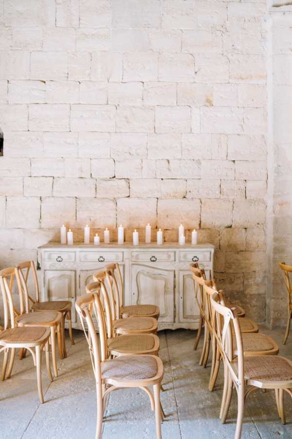 A ceremony setup inside a stone-walled interior space, likely a historic chateau or cave venue in France. Natural wood cross-back chairs with woven rattan seats are arranged in rows facing a distressed grey-painted wooden sideboard, which is lined with varying heights of white pillar candles as the sole decorative element. The decor palette is minimal and neutral, relying on warm wood tones, off-white candles, and the pale limestone walls and stone-tiled floor. Wide shot capturing the full ceremony seating arrangement and altar area. Potential venue feature image.