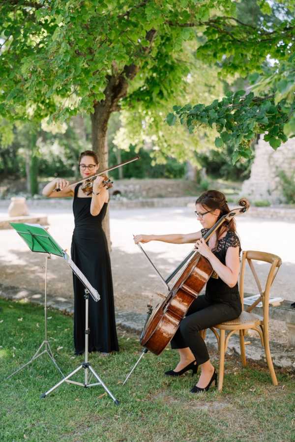 Two female musicians perform outdoors at a wedding, one standing and playing violin while the other sits on a natural wood cross-back chair playing cello. Both are dressed in black — the violinist in a long black sleeveless gown and the cellist in a black lace top and trousers. A green music stand holds their sheet music in the foreground. The setting is an outdoor garden or grounds area with a gravel pathway visible in the background, consistent with a French chateau or abbey property. The shot is a medium portrait-style image taken in natural daylight.