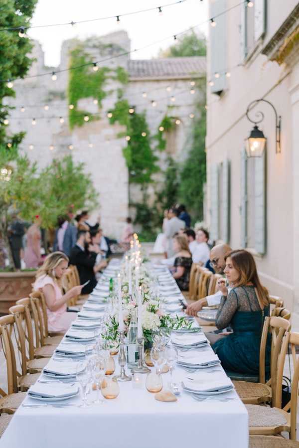 An outdoor wedding reception dinner is underway in a courtyard between a white rendered building and an old stone wall covered in climbing greenery. Approximately 15–20 guests are seated along a single long banquet table dressed in a white linen tablecloth, with grey linen napkins, white dinner plates, and amber-tinted glassware. The centerpiece runs the length of the table and features clusters of white hydrangeas, blush florals, and trailing greenery interspersed with tall white taper candles in glass holders. Natural wood cross-back chairs line both sides of the table. Overhead, warm globe string lights are strung across the courtyard, providing the primary ambient lighting as the evening light fades. Guests are dressed in a mix of formal attire including a teal lace dress, blush pink dress, and dark suits. The overall styling is classic French with a relaxed, intimate feel. Wide shot taken from one end of the table looking down its full length. Potential venue feature image.