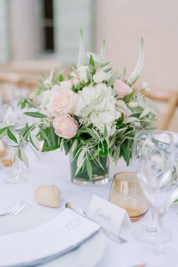 Close-up detail shot of a wedding reception table setting featuring a low centerpiece arrangement of blush pink garden roses, white hydrangeas, white lisianthus, and trailing olive branch greenery in a clear glass vase. The table is dressed with a white linen, white charger plates, silver cutlery, a bronze-toned tumbler glass, crystal wine glasses, and a small white calligraphy place card reading 'Camille' alongside a stone-colored favor. The overall decor palette is white, blush pink, and soft green with a classic, clean aesthetic. The background is softly blurred, suggesting an indoor or covered outdoor reception space.