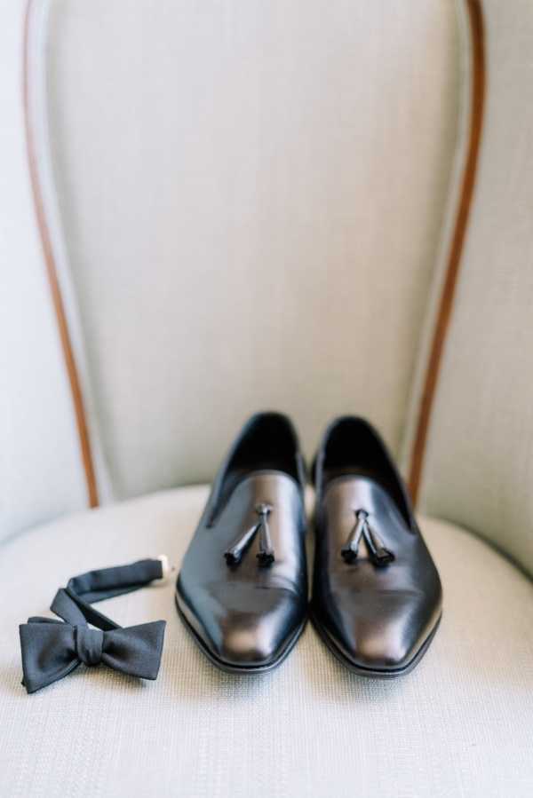 A close-up detail shot of groom's accessories arranged on a cream upholstered chair. A pair of black leather tassel loafers are placed side by side on the seat, with a charcoal grey satin bow tie laid to the left of them. The composition is a styled flat-lay-style portrait taken from a slightly elevated angle, with the chair back providing a soft, neutral background. The overall styling suggests a classic, formal aesthetic.