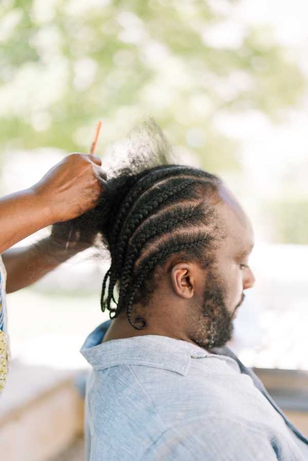 A getting-ready scene showing a man having his braided hair styled, with a hand holding his cornrow braids upward and a comb visible. The man, seen in three-quarter profile, has neat cornrows along the sides of his head and a short beard, and is wearing a light blue chambray shirt. The shot is taken outdoors or on a covered terrace in natural diffused light, with a softly blurred green background. This is a close-up portrait-style composition focusing on the hair styling preparation.