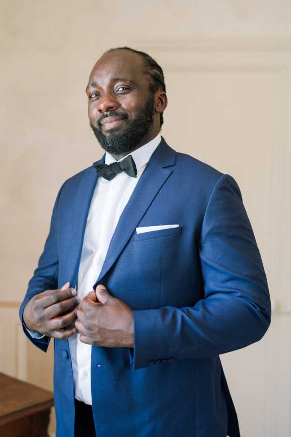 A getting-ready portrait of the groom indoors, photographed against a light neutral wall. He is wearing a navy blue suit jacket with a white dress shirt, black bow tie, and a white pocket square, and is in the process of buttoning his jacket. The shot is a close-up portrait framing him from roughly the waist up, with natural light creating soft, even tones on his face.
