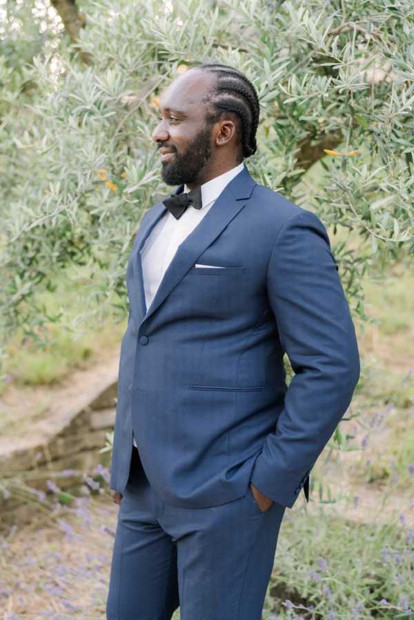A portrait of the groom standing outdoors, shot from approximately mid-thigh up. He wears a navy blue suit with a white dress shirt, a black bow tie, and a white pocket square. His hair is styled in cornrow braids and he has a neatly groomed beard; he is looking to one side with his hands in his pockets. The background features olive tree foliage and a hint of lavender plants at ground level, suggesting a French countryside or Provençal venue setting.