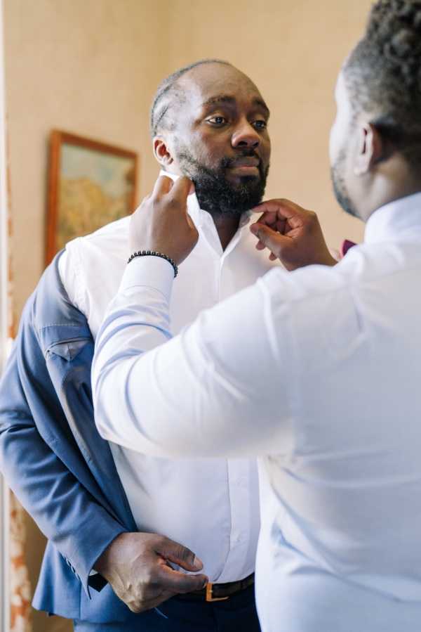 A getting-ready moment captured indoors, showing two men — likely the groom and a groomsman or family member — as one helps the other adjust his collar and shirt. The groom wears a white dress shirt, navy blue trousers with a tan belt, and a light blue suit jacket partially on, along with a black beaded bracelet. The person assisting wears a white shirt and appears to have a burgundy accessory at his collar. The setting is a warm-toned interior room with a framed painting visible on the wall in the background. The shot is a close-up portrait taken from a slightly over-the-shoulder perspective of the helper, keeping focus on the groom's face and upper body.