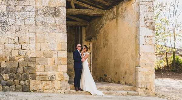 A couple poses together for a portrait in the shaded archway of a large stone structure, likely a historic chateau or fortified building with rough-cut limestone block walls and a visible wooden beam ceiling overhead. The bride wears a slim, floor-length white gown with a simple silhouette, while the groom is dressed in a navy suit and sunglasses, standing behind her with his arms around her waist. The shot is a medium-wide portrait capturing both figures full-length within the architectural frame. The setting has a rustic, historic character with weathered stone surfaces and an outdoor courtyard visible to the right.