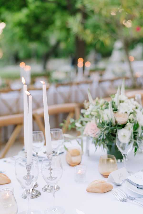 Close-up detail shot of an outdoor wedding reception table set with a white linen tablecloth, two lit white taper candles in silver candlestick holders, and crystal glassware including wine glasses filled with white wine. Small bread rolls are placed at each setting alongside silver cutlery and white place cards. A low centerpiece arrangement features blush pink roses, white blooms, and lush greenery. In the background, additional round tables are set with matching decor, and natural wood cross-back chairs are visible, along with warm string lights glowing softly out of focus. The overall decor palette is white, blush, and silver with a classic, garden-style aesthetic.