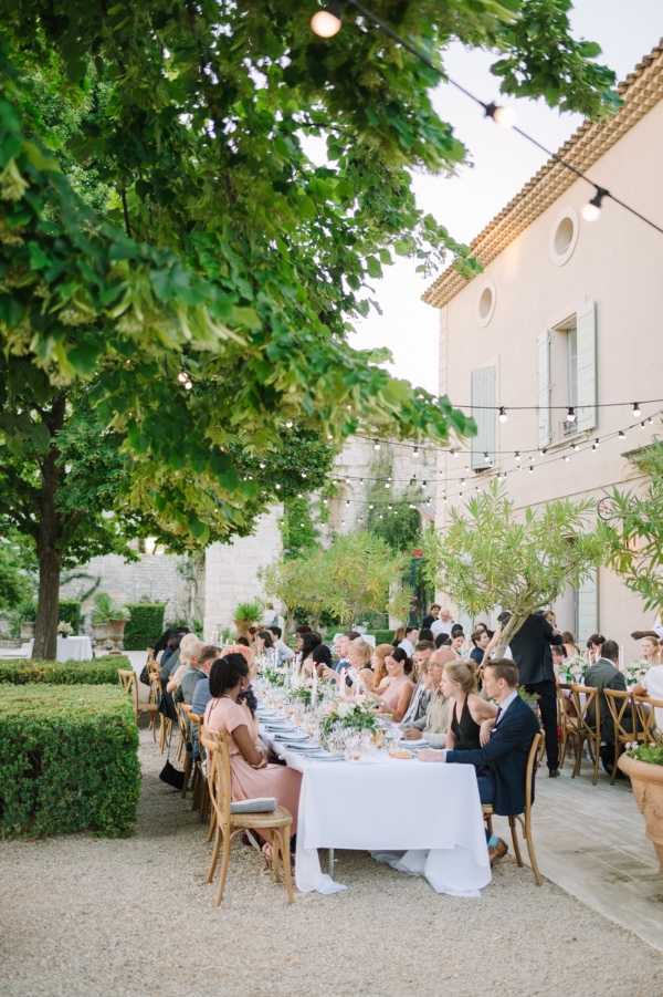 An outdoor wedding reception dinner is taking place in the courtyard of a French mas or bastide-style property, with stone and rendered walls, terracotta roof tiles, and pale green shutters visible in the background. Approximately 25–30 guests are seated along a long rectangular table dressed in a white linen tablecloth with slate blue napkins, low floral centerpieces in white and greenery, and glassware. Guests are dressed in a mix of smart casual attire including a dusty pink midi dress and a navy suit visible in the foreground. Warm bistro string lights are strung overhead between the building and surrounding trees, and the setting is styled in a relaxed, classic Provençal aesthetic with natural wood cross-back chairs and a gravel courtyard. The shot is a medium wide angle taken from a slight elevation, framed by an overhanging tree canopy in the upper left. Potential venue feature image.