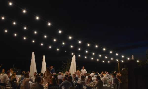An outdoor wedding reception dinner is taking place at night, with a large group of guests — approximately 60 or more — seated at round tables arranged across an open terrace or garden. The tables are dressed in white linens and set with glassware, and white closed patio umbrellas are visible in the background. The scene is lit entirely by strands of Edison-style festoon bulb lights strung overhead in parallel lines, creating a warm, ambient glow across the space. The wide shot captures the full reception layout from a slightly elevated angle, with a staff member in white visible standing among the tables.