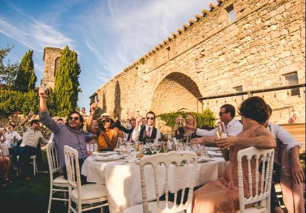 An outdoor wedding reception dinner is taking place in the courtyard of a historic stone building with a large arched opening in the wall, likely a French chateau or abbey. Approximately 8-10 guests are visible in the foreground seated around a round table dressed in white linen with glassware and place settings, raising their glasses in a toast, with additional guests visible at tables in the background. The guests are dressed in casual-to-smart summer attire in warm tones including mustard yellow, blush pink, and light blue. White Napoleon/Chiavari chairs surround the tables, and the overall decor style is relaxed and rustic, suited to the historic stone venue. The image is a wide shot taken in bright afternoon sunlight.