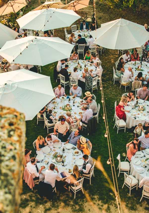 An aerial shot of an outdoor wedding reception dinner taking place on a garden lawn, with approximately 60-80 guests seated at multiple round tables covered in white linens. The tables are arranged across the grass and shaded by large white market umbrellas, with white Chiavari chairs surrounding each table. A string of exposed bulb bistro lights runs diagonally across the scene, suggesting evening ambiance as the light appears to be late afternoon golden hour. Guests are dressed in smart-casual summer attire, with pops of red, yellow, and coral visible among the crowd. Table settings appear to include white plates, wine bottles, and minimal greenery centerpieces. The shot is taken from an elevated position, likely from a terrace or upper floor of a nearby building, giving a full overhead view of the reception layout.