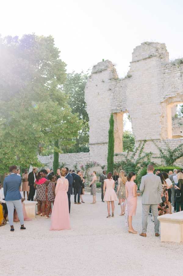 Cocktail hour taking place outdoors in the courtyard of a historic ruined stone structure, likely in the South of France, with tall arched openings and partial walls integrated with climbing greenery and cypress trees. Approximately 30 guests are mingling on a gravel surface, dressed in a mix of smart-casual and formal attire including blush pink full-length and midi dresses, a light blue suit, and a pink hat. The warm golden light suggests late afternoon or early evening, giving the scene a sun-drenched, summery feel. Wide shot taken from behind the crowd, capturing the full architectural backdrop of the ruins. Potential venue feature image.