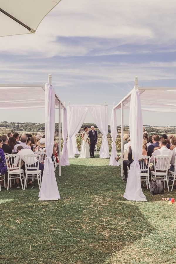 An outdoor civil or symbolic ceremony is taking place on a lawn with an open countryside view in the background. The couple stands at the far end of a central aisle beneath a white fabric arch structure, with the bride in a white gown holding a bouquet and the groom in a dark suit. Guests — approximately 40-50 people — are seated on white Napoléon chairs arranged in two rows on either side of the aisle, which is flanked by white pillar structures draped with flowing pale lavender fabric tied at the base. The overall decor palette is white and soft lavender, giving a clean, minimal aesthetic. The shot is taken from the back of the aisle at ground level, providing a wide perspective that frames the couple through the fabric-draped columns.