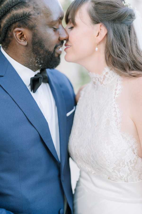 Close-up portrait of a bride and groom leaning in for a kiss, shot with a shallow depth of field that softly blurs the background. The groom wears a navy blue suit jacket with a white dress shirt and black bow tie, while the bride wears an ivory halter-neck lace gown with a high neckline and pearl drop earrings, her dark hair styled in a half-up arrangement with a small floral hair accessory. The composition is tightly framed on their faces and upper torsos, emphasizing the intimate moment between the couple.