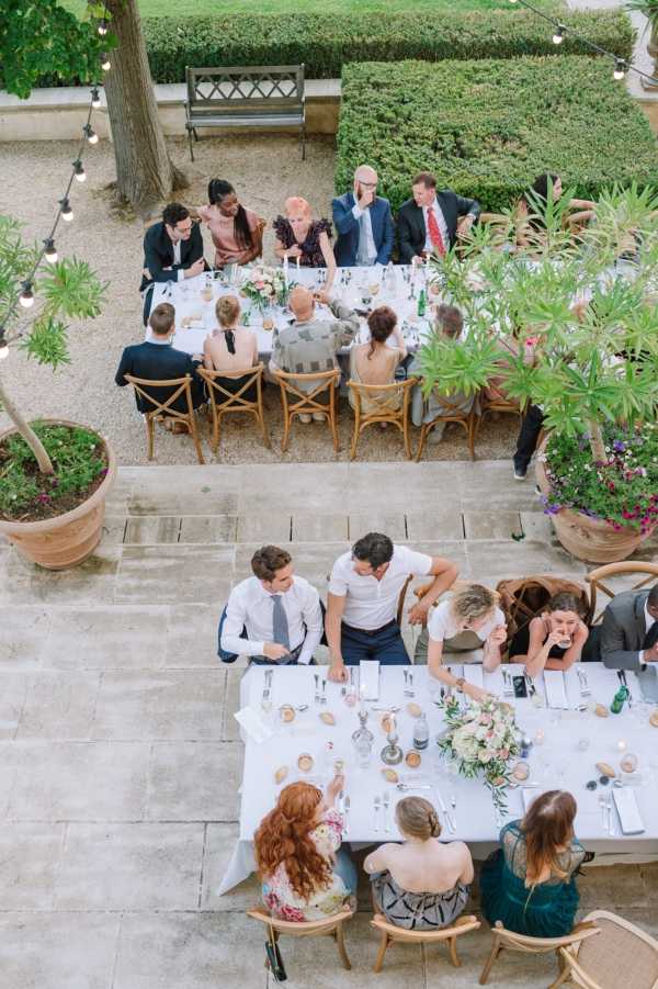 An aerial shot of an outdoor wedding reception dinner taking place on a stone terrace, with approximately 20 guests seated across two long rectangular tables. The tables are dressed with white linen cloths, silver cutlery, and low floral centerpieces featuring blush and ivory blooms with greenery. Cross-back wooden chairs are used throughout. String Edison bulb lights are strung overhead, and large terracotta planters with green shrubs and flowering plants frame the space. Guests are dressed in a mix of smart casual and semi-formal attire, including a teal velvet dress, a floral print dress, and suits in navy and grey. Green wine bottles are visible on the upper table. The setting has a classic French garden aesthetic with trimmed hedgerows and a stone bench visible in the background.