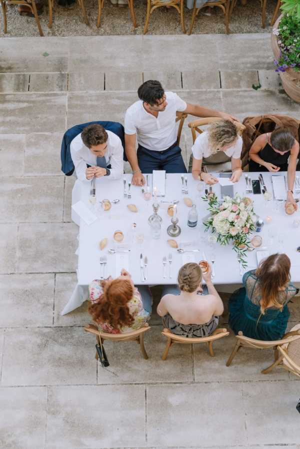 Aerial shot of a wedding reception dinner table set on an outdoor stone-paved courtyard or terrace. Seven guests are seated around a rectangular table covered with a white linen tablecloth, engaged in conversation. The table is styled with a central floral arrangement of ivory and blush pink flowers with olive or eucalyptus greenery, small votive candles, glass decanters, silver cutlery, folded white napkins, and blush-toned glassware. Guests are dressed in a mix of smart casual attire including a navy suit jacket, a teal lace dress, a floral dress, and a dark strappy top. Natural wood cross-back chairs are visible at the top of the frame, suggesting additional seating in the background.