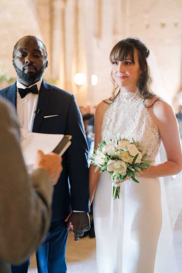 A mid-shot of a bride and groom standing together during an indoor ceremony, facing an officiant who is partially visible in the foreground holding papers. The bride wears a white sleeveless halter-neck gown with a lace bodice and a flowing skirt, paired with a cathedral-length veil and bangs with her hair loosely pinned up; she holds a compact bouquet of white roses and garden roses with green foliage. The groom stands beside her in a navy suit with a white dress shirt and black bow tie. The setting appears to be a stone-walled venue, possibly a chapel or historic chateau hall, with warm ambient lighting visible in the background.