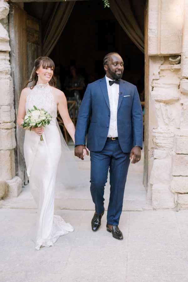 The bride and groom walk hand-in-hand through a large stone archway doorway, likely just after their ceremony at a chateau or historic stone venue in France. The bride wears a sleek ivory halter-neck gown with a lace bodice and a flowing skirt with a small train, carrying a bouquet of white roses and green eucalyptus. The groom wears a navy blue suit with a white waistcoat, black bow tie, and black tassel loafers. This is a mid-length portrait shot capturing the couple exiting the venue entrance, with guests and draped fabric visible in the darkened interior behind them.