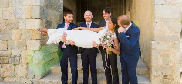 A fun group portrait taken outdoors in front of a stone building, showing the groom and three groomsmen lifting and carrying the bride horizontally while she holds her bouquet. The groom and groomsmen are all dressed in navy suits with light pink ties, while the bride wears a white lace wedding dress. The bride holds a loose, garden-style bouquet featuring pink and coral blooms with greenery. The group is laughing and smiling, creating a playful mood. The shot is a medium wide portrait framing all five people against the rustic stone wall backdrop.