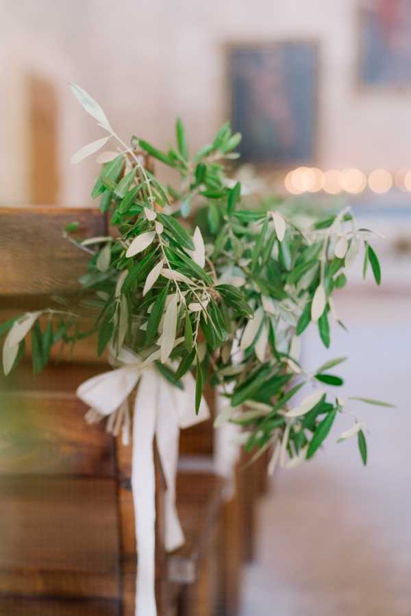 Close-up detail shot of a wooden church pew decorated with a sprig of olive branches tied with a trailing ivory satin ribbon, used as aisle decor for a wedding ceremony. The greenery-only styling with no blooms creates a minimal, nature-forward aesthetic. In the soft-focus background, warm candlelight and what appears to be a painting or artwork on a light-colored interior wall are visible, suggesting an indoor chapel or historic venue setting. The overall decor palette is green and ivory with warm ambient lighting.