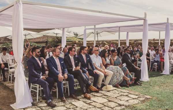 An outdoor wedding ceremony in progress, with approximately 40–50 guests seated in white chairs beneath a large white fabric canopy structure with draped curtain panels at the posts. The setting appears to be a rural French property with low stone walls and terracotta-roofed buildings visible in the background, along with large white parasols providing additional shade. Guests in the foreground include several men in navy and charcoal suits with light pink and white ties, and women in floral and patterned summer dresses. The overall decor style is clean and classic with an all-white canopy palette. The shot is a wide-angle view taken from the side of the seating area, capturing the crowd's attention directed toward an unseen officiant or couple at the front.