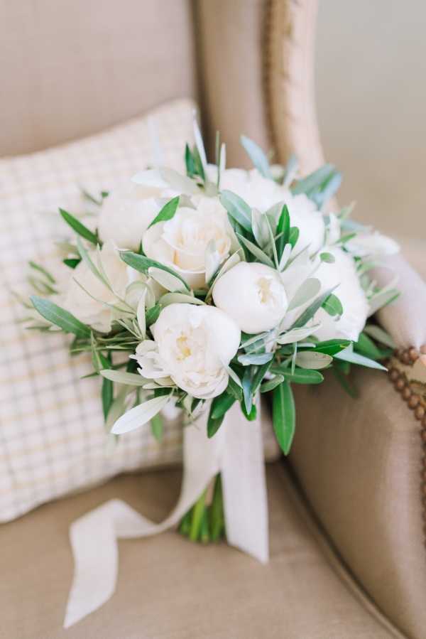 Close-up detail shot of a bridal bouquet resting on a beige upholstered armchair with a small checked cushion behind it. The bouquet is composed of white peonies and olive branch foliage with silver-green leaves, tied with a trailing ivory silk ribbon. The arrangement is compact and rounded, with a classic, garden-inspired aesthetic in a white and green palette.
