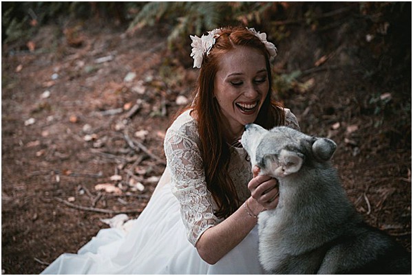 A bride sits on the ground in an outdoor woodland setting, laughing joyfully as a grey and white Husky puppy nuzzles or licks her hand. She wears a white lace long-sleeve wedding dress and has a small white floral hair accessory in her long red hair. The portrait-style shot is taken at ground level, with a natural forest floor of pine needles and dirt in the background. The styling is boho with the lace dress detail and floral headpiece.