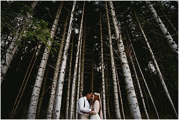 A couple portrait taken outdoors in a dense forest setting, with the bride and groom standing close together and leaning in for a near-kiss at the base of a cluster of tall, pale-trunked birch trees. The groom is dressed in a white shirt with dark suspenders, while the bride wears a long-sleeved, flowing ivory or cream dress and a floral crown with small blooms in her hair. The composition is a wide shot that uses the vertical lines of the tree trunks to frame the couple centrally, with the canopy creating a dark, moody backdrop that contrasts with the lighter trunks. The overall styling has a boho aesthetic, and the image has a dark, atmospheric quality with the couple lit as a focal point against the shadowy forest.